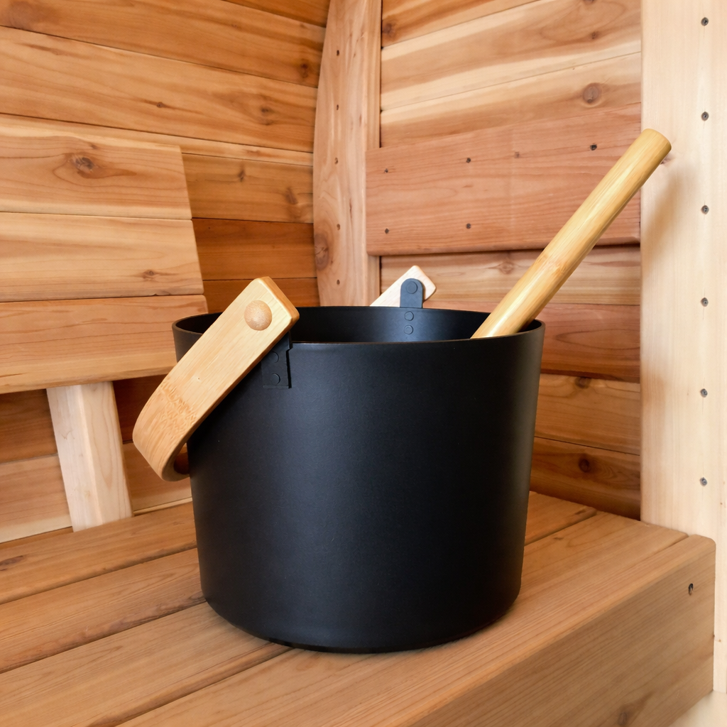 Modern black metal sauna bucket and ladle placed on a cedar bench inside a red cedar barrel sauna interior