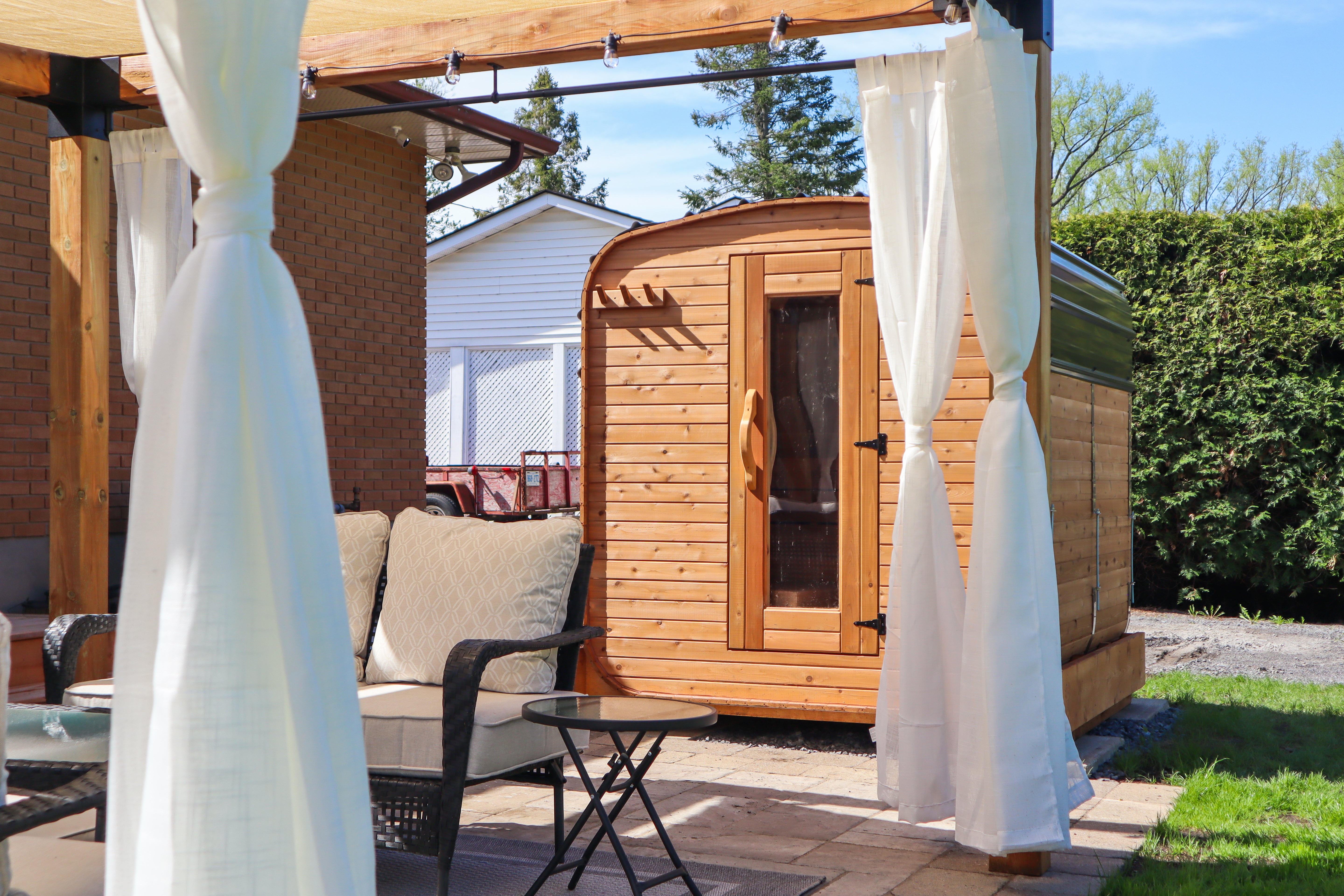 Cube cedar outdoor sauna beside a backyard seating area and gazebo, modern patio wellness setup in a landscaped residential yard