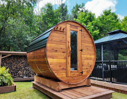 6-ft red cedar barrel outdoor sauna on a backyard wood deck platform with stacked firewood beside the unit, cozy residential sauna setting