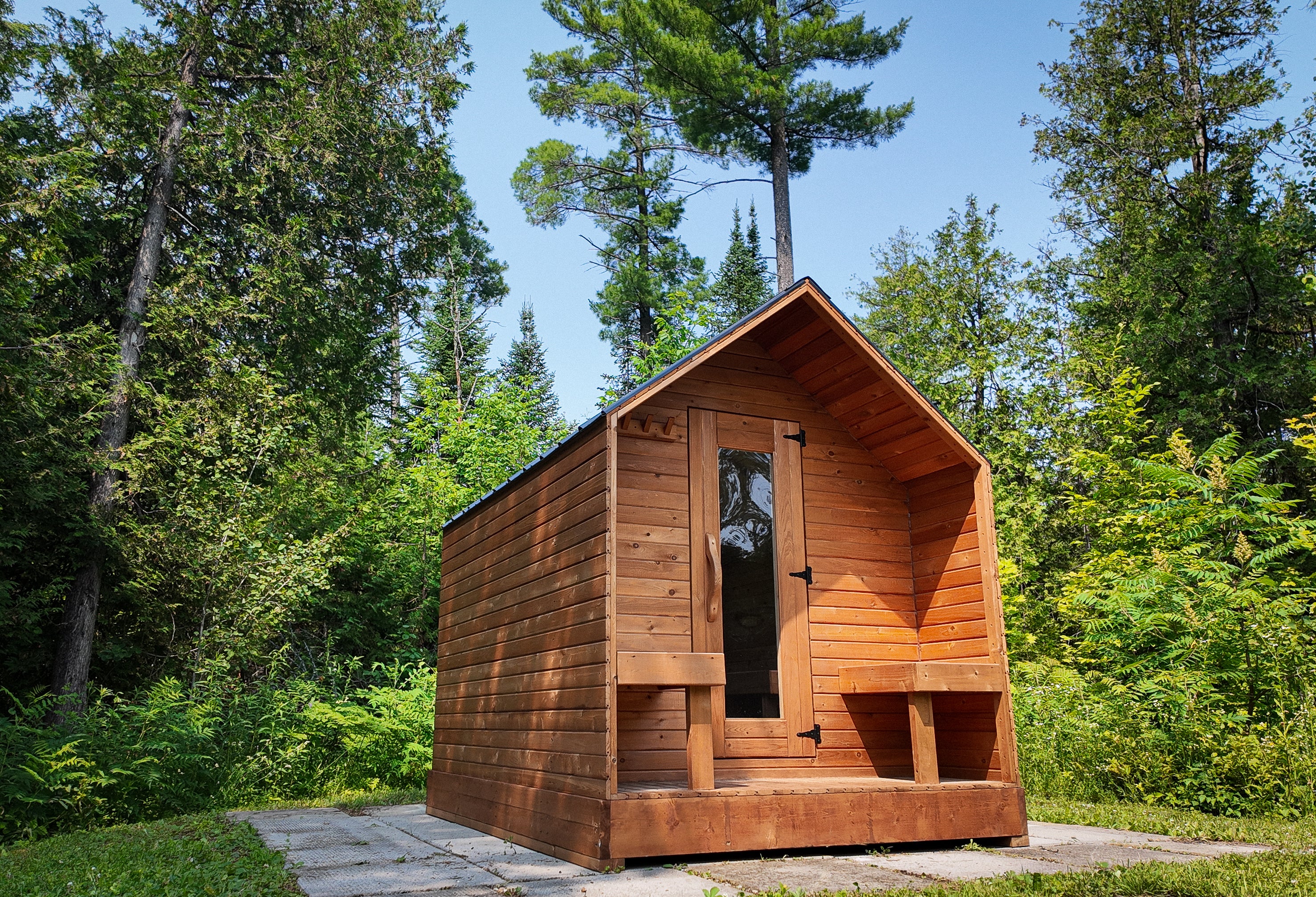 Front view of real red cedar cabin outdoor sauna with covered porch and glass door entrance in a wooded backyard landscape
