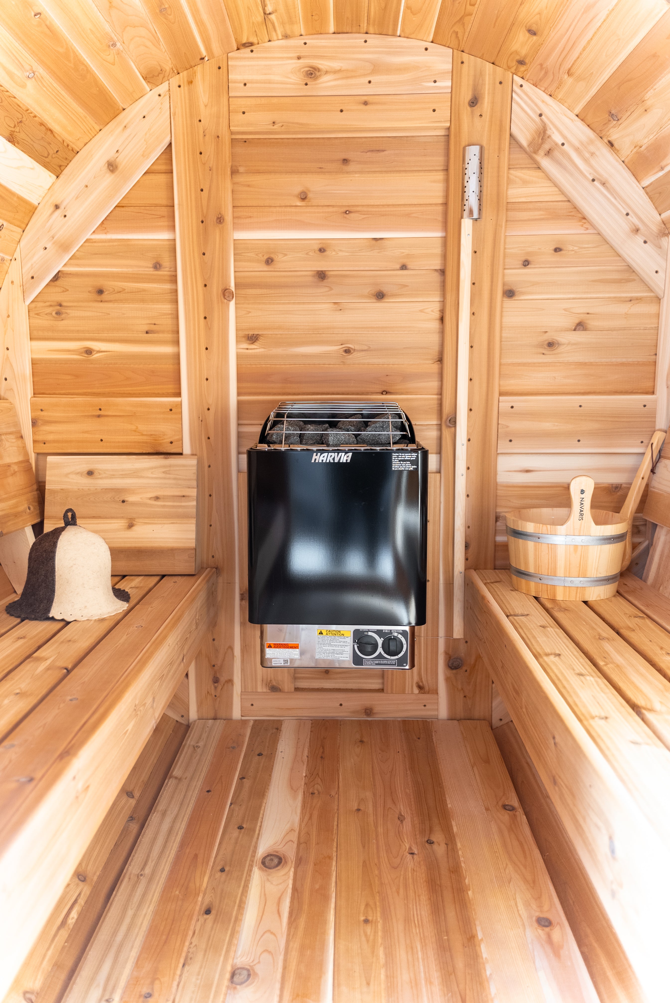 Harvia KIP electric sauna stove wall-mounted inside a cedar barrel sauna, with a wooden bucket, ladle, and sauna hat placed on either side