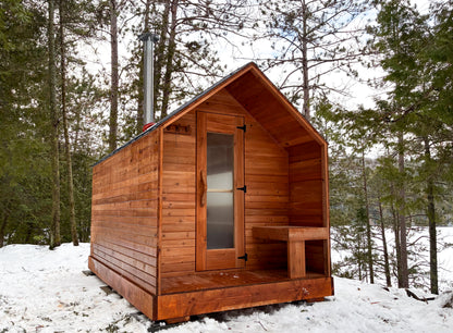 Front view of real red cedar cabin outdoor sauna with covered porch and glass door entrance in a wooded cottage landscape
