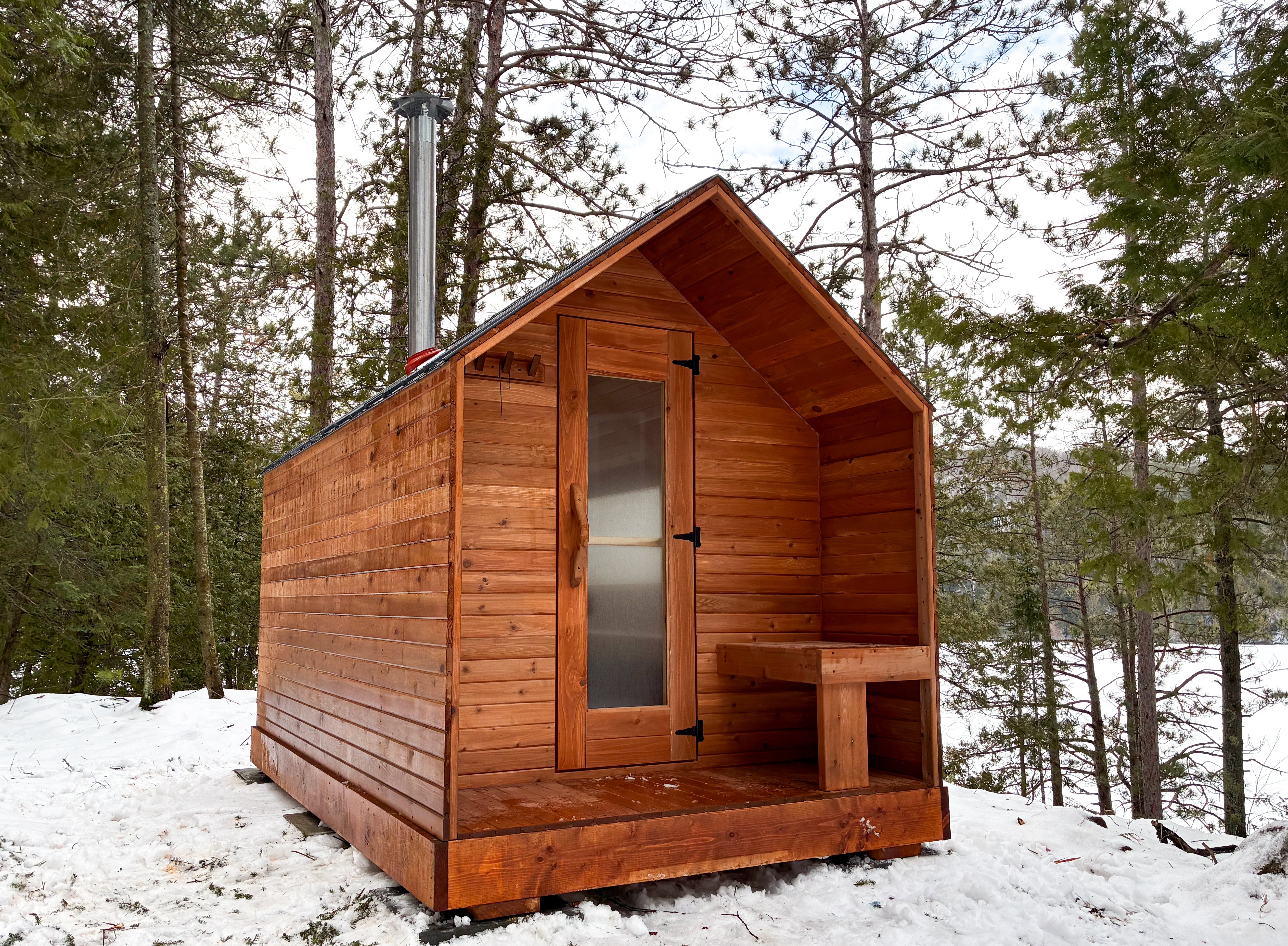 Front view of real red cedar cabin outdoor sauna with covered porch and glass door entrance in a wooded cottage landscape