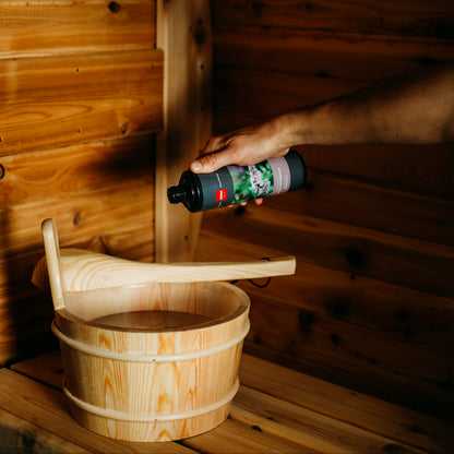 Hand pouring sauna essential oil scent into a wooden bucket inside a cedar sauna interior